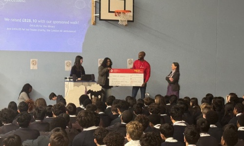House captains handing over a large cheque to London Air Ambulance representative, in front of the Lamb students during their assembly